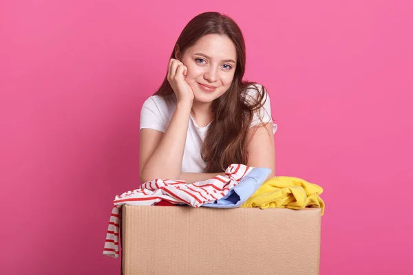 Donation Concept. Woman holding donate box with full of clothes, adorable smiling female keeps hand under chin and clothes donate box, lady wearing casual white t shirt, posing isolated on pink. — Stock Photo, Image