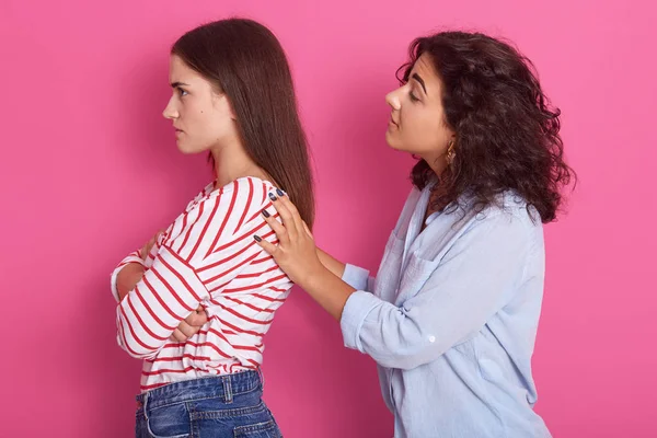 Beautiful slim Caucasian ladies wearing casual clothing, do not talk to each other, woman dressed blue shirt touches female's shoulders, models posing isolated over pink wall. Relationship concept. — ストック写真