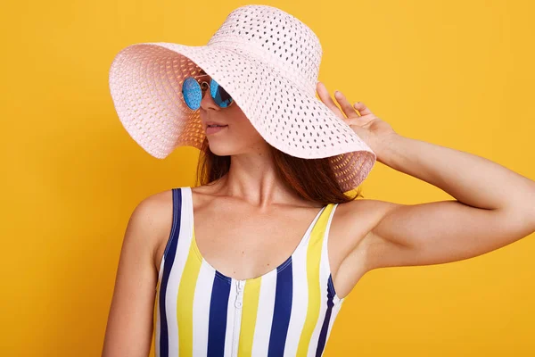 Close up portrait of beautiful girl wearing striped colored swimming suit, hat and sun glasses, adorable female tounching her cap and looking aside, posing isolated over bright yellow background. — ストック写真