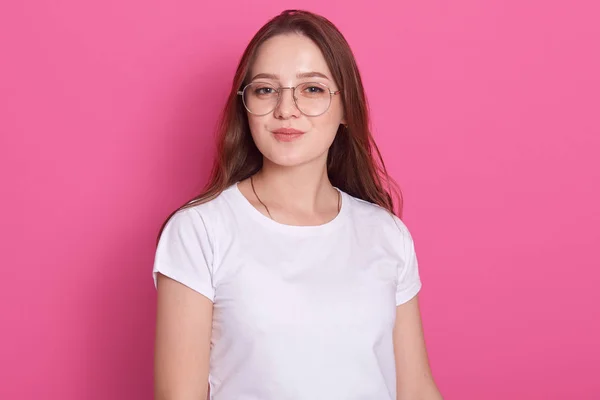 Primer plano de la joven estudiante atractiva feliz con el pelo largo staraight, vistiendo camiseta casual blanca y gafas, atractivos puestos femeninos sonriendo aislado sobre fondo rosa. Concepto de personas . — Foto de Stock