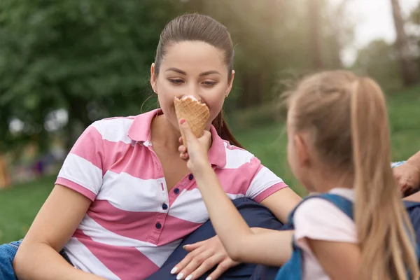 Close up image of young happy family spending their weekend in park, relaxing on frameless chair, charming daughter feeding her moomy with ice cream, enjoing to spend time together, erxpressing joy. — Stock Photo, Image