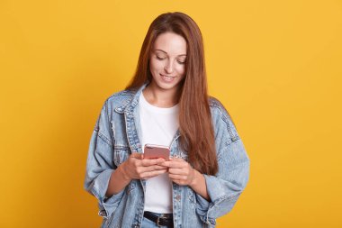 Koyu renk düz saçlı güzel bir kadının portresi. Telefonuna mesaj atıp gülümsüyor, sosyal ağlara bakıyor. Kot ceket giyiyor, sarı arka planda izole edilmiş poz veriyor..