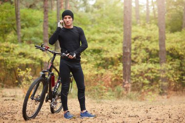 Siyah eşofman ve spor ayakkabı giyen, dağ bisikletinin yanında duran ve ormanlık alanda telefonla konuşan, aktif bir şekilde zaman geçiren bir adamın açık hava fotoğrafı. Sağlıklı yaşam tarzı kavramı.