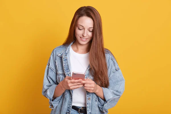 Potret wanita muda cantik yang bahagia dengan pesan SMS rambut lurus gelap di ponselnya dan tersenyum, memeriksa jaringan sosial, mengenakan jaket denim, berpose terisolasi di atas latar belakang kuning . — Stok Foto