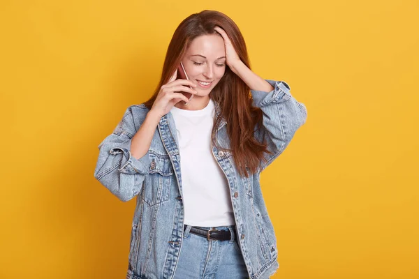 Portret van tevreden goed uitziende vrouw jurken modieuze spijkerjasje en wit casual shirt, holding smartphone, het hebben van een gesprek, houdt de hand op haar hoofd, geïsoleerd over gele achtergrond. — Stockfoto