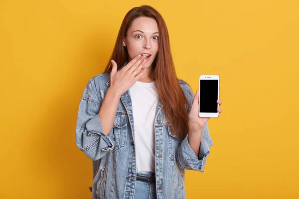 Close up portrait of young beautiful shocked woman dresses casual style, holding phone with blank screen and keeps palm on her mouth, looking at camera. Studio shot isolated over yellow background. — Stock Photo, Image