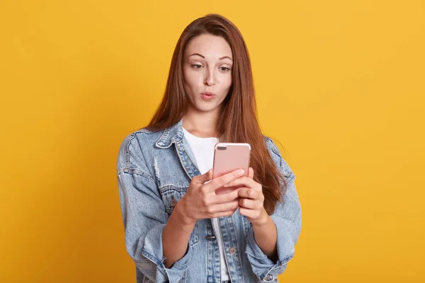 Portrait of attractive Cauacasian woman gets surprising news via telephone call, keeps lips rounded, wears stylish denim jacket, has eyes popped out, hears astonishing relevation, standing indoor. — ストック写真