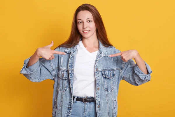 Portrait of beautiful girl with dark straight hair and no makeup, dress denim jacket and white shirt, stands pointing himself with both hands and look at camera, isolated on yellow background . — Stok Foto
