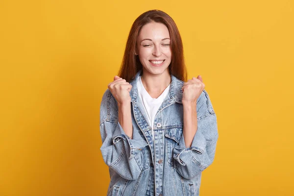 Disparo horizontal de feliz mujer lograda y alegre celebrando la victoria o el éxito, posando y apretando los puños en triunfo, mantiene los ojos cerrados, mira sonriendo a la cámara aislada sobre el fondo amarillo —  Fotos de Stock