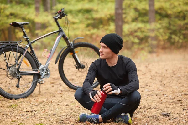 Man wearing black track suit and cap sitting onground with crossd legs near his bicycle, cyclist stops to drink water, holding red bottle in hands, looks thoughtfully aside, having rest in forest.