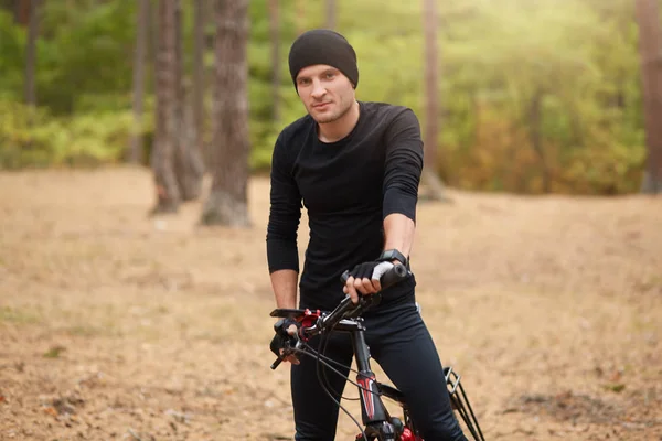 Cyklista na horských kolech na jedné trati venkovní se stromy na pozadí, atraktivní muž v černém sportovním oblečení a čepici stojící s bicyklem v lese, tráví volný čas. Sportovní koncept. — Stock fotografie