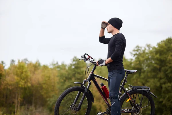 Beeld van road biker fietsen en training op de weg in het bos. Aantrekkelijke jonge sporter stopt om te rusten na lange uren rijden, houdt de hand dicht bij het voorhoofd en kijkt ver weg. Gezond activiteitsconcept. — Stockfoto