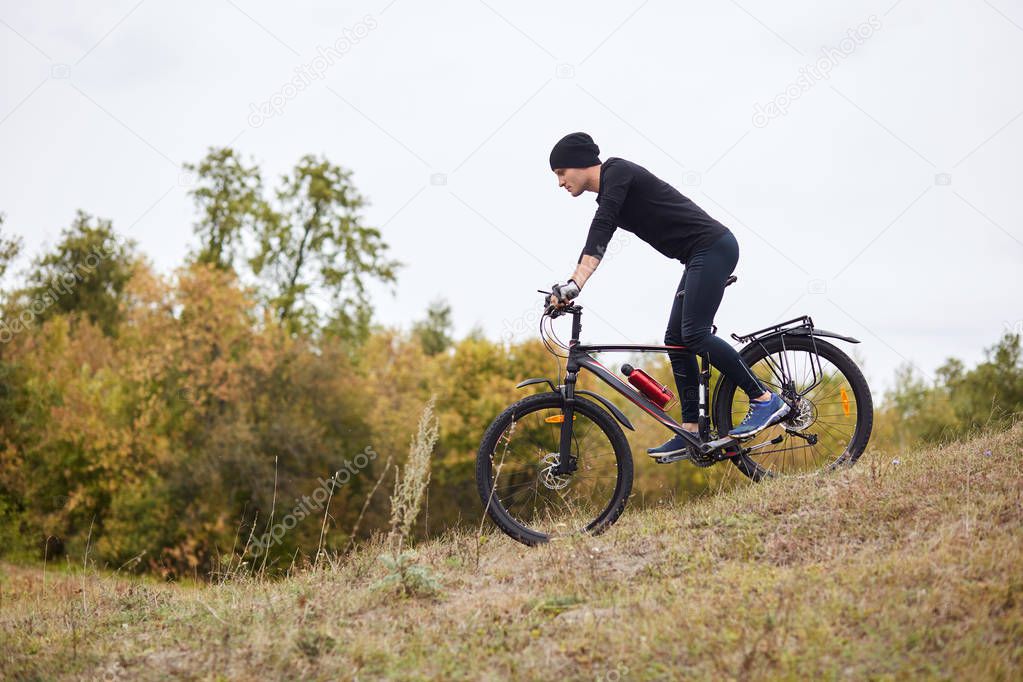 Side view of sporty man riding mountain bike on enduro track, guy dresses black track suit and cap, ridiing down hiil in forest, being very concentrated, engoing road cycling and mountain biking.