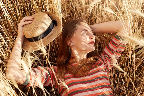 Modelo magnético relaxado deitado no campo de trigo, desfrutando do tempo em torno da natureza, escondendo-se do sol, tirando seu chapéu de palha, segurando-o em uma mão, olhando para o lado, estando em espíritos elevados. Conceito de natureza . — Fotografia de Stock