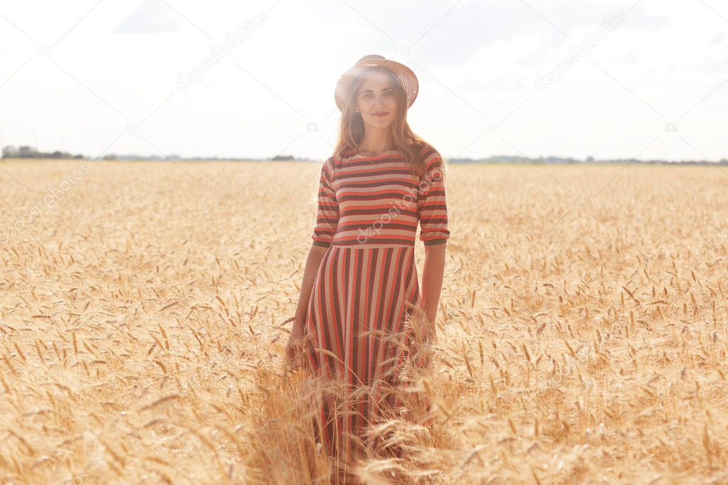 Cheerful attractive young woman standing straight in filed at rural area, feeling nature, being inspired with landscape, having pleasant facial expression, wearing casual clothes. Emotions concept