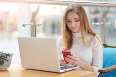 İnternette çalışan, kafede oturan, dizüstü bilgisayarını ve cep telefonunu aynı anda kullanan, uzun sarı saçlı ve görevlere odaklanmış ciddi meşgul bir genç kız görüntüsü. İnsanlar ve iş zamanı kavramı.