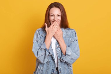 İki eliyle ağzını kapatan, utangaç, şaşkın bakışlı, neşeli, kot ceket ve beyaz t-shirt giyen, stüdyoda sarı arka planda izole edilmiş genç bir model..