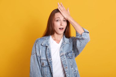 Duygusal açıdan tatlı, güzel bir genç ağzını açıyor, bir elini alnına koyuyor, şaşırmış hissediyor, doğrudan kameraya bakıyor, stüdyoda sarı arka planda tek başına duruyor..