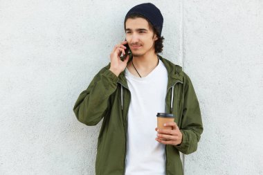 İnce, hoş bir çocuk yana bakıyor, akıllı telefonunu kulağının dibinde tutuyor, konuşmaya odaklanmış, beyaz tişört, yeşil ceket ve siyah şapka giyiyor, elinde kağıt bardak kahve tutuyor. Gençlik ve zaman kavramı.