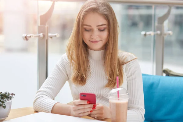Kırmızı akıllı telefonunu elinde tutan, nazikçe gülümseyen, sosyal ağ sitelerini kullanan, kafede tek başına oturan, masasında kuyruk kuyruğu olan, keyifli bir kız. Teknoloji kavramı. — Stok fotoğraf
