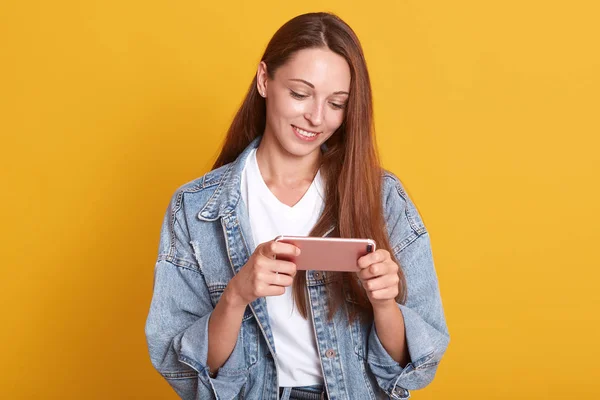 Image of tender attractive girl smiling sincerely, holding pink smartphone in her hands, looking at its screen, watching video, being fond of new technologies, wearing trendy clothes. Tech concept. — Stock Photo, Image