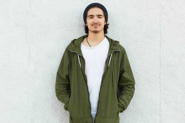 Image d'un jeune homme calme enchanté debout à l'extérieur, ayant une expression faciale agréable. souriant calmement, regardant directement la caméra, mettant les mains dans les poches. Concept de style rue . — Photo