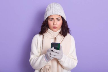 Akıllı telefonundaki mesajlarla ilgilenen sevimli genç bir kız, kötü haberleri okuyor, yüz ifadesini hayrete düşürüyor, kalın kazak, kep, atkı ve eldiven giyiyor, leylak duvarda izole edilmiş..