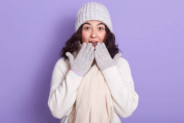 Молодая привлекательная девушка в белом свитере, теплая кепка, шарф и перчатки, с легким макияжем, закрывая рот руками, глядя с большими глазами в удивлении, изолированные на сиреневом фоне . — стоковое фото