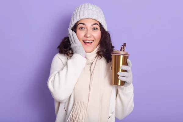 Joven mujer hermosa posando con sonrisa y expresión facial sorprendida, bebiendo té de la taza termo, moviéndose hacia cero residuos, mantiene la palma en su mejilla, posando con la boca abierta, lleva ropa de abrigo . — Foto de Stock