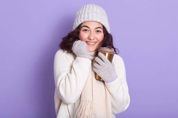 Νεαρή attarctive γυναίκα basking τα χέρια της μέσω φλιτζάνι ζεστό ρόφημα και γάντια, φορώντας πουλόβερ, κασκόλ και καπέλο, θέτοντας απομονώνονται σε μωβ φόντο, χρησιμοποιώντας κούπα θερμό, κινείται προς το μηδέν απόβλητα. — Φωτογραφία Αρχείου