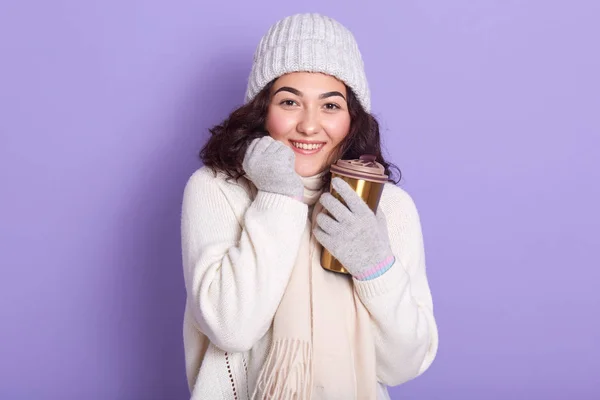 Ung attarctive kvinna sola händerna via kopp varm dryck och handskar, bär tröja, halsduk och mössa, poserar isolerad över lila bakgrund, med hjälp av termo mugg, rör sig mot noll avfall. — Stockfoto