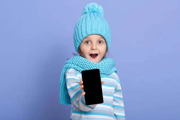 Petite fille étonnante enfant portant un chapeau d'hiver avec un pompon montrant l'écran vide pour l'espace de texte du nouveau téléphone mobile populaire isolé sur fond bleu, enfant debout avec la bouche ouverte et surpris — Photo