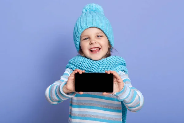 Souriant petite fille enfant robes chapeau d'hiver moderne avec pompon, écharpe et chemise décontractée rayée, montrant écran vide de téléphone intelligent, charmant modèle debout isolé sur fond bleu. Espace de copie . — Photo