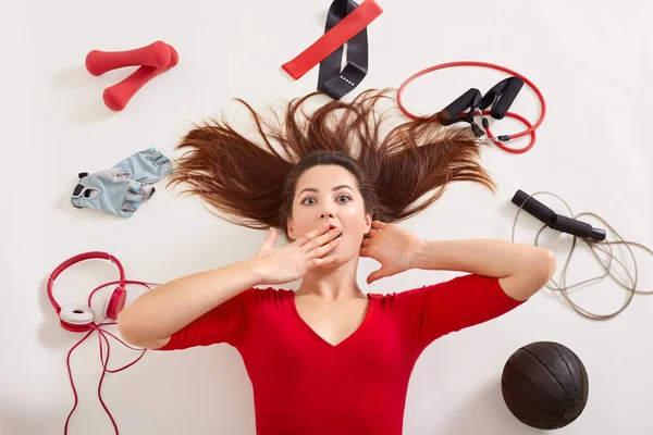 Estudio de mujer caucásica sorprendida tendida en superficie blanca con expresión facial asombrada, cubriéndose la boca con la mano, rodeada de equipos de esportismo: expansor, guantes de fitness, bandas elásticas . — Foto de Stock