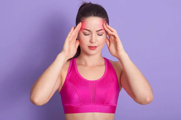 Atractiva deportista con terrible dolor de cabeza tocando sus sienes con ambas manos, mantiene los ojos cerrados, posando aislada sobre fondo de estudio lila, chica deportiva con top rosa. Concepto de cuidado saludable . — Foto de Stock