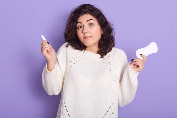 Foto de atractiva mujer morena sostiene tampón de algodón y servilleta sanitaria, que tiene ciclo menstrual mensual, lleva camisa blanca, no sabe que para elegir por el período, isoalted sobre fondo púrpura . — Foto de Stock