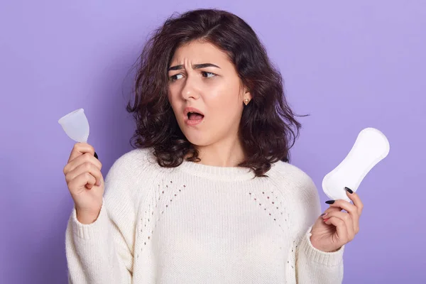 Estudio de tiro de mujer sosteniendo copa menstrual y sanitarypad en las manos sobre fondo lila brillante, no sabe qué elegir, se levanta con la boca abierta. Producto de higiene femenina durante el concepto de período . — Foto de Stock