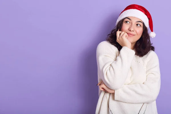 Задумчивая юная брюнетка смотрит в сторону, трогает подбородок рукой, носит белый свитер и красную шляпу Санта Клауса, ждет новогодней вечеринки, радуется. Копирайт для рекламы . — стоковое фото