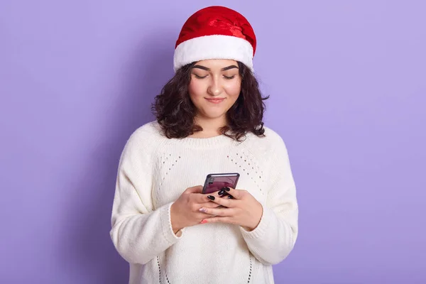 Изображение привлекательной красивой молодой женщины, держащей в руках смартфон, пишущей поздравления с Новым годом, в белом свитере и красной шляпе Санта Клауса, изолированной на фоне сирени в студии . — стоковое фото