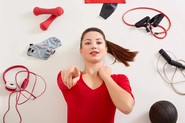 Wanita muda menggemaskan memegang titik di kamera dengan jari telunjuknya saat berbaring di permukaan putih, gadis dengan gaun ekor kuda pakaian merah, headphone, expander, dumbbels, band elastis, sarung tangan kebugaran — Stok Foto