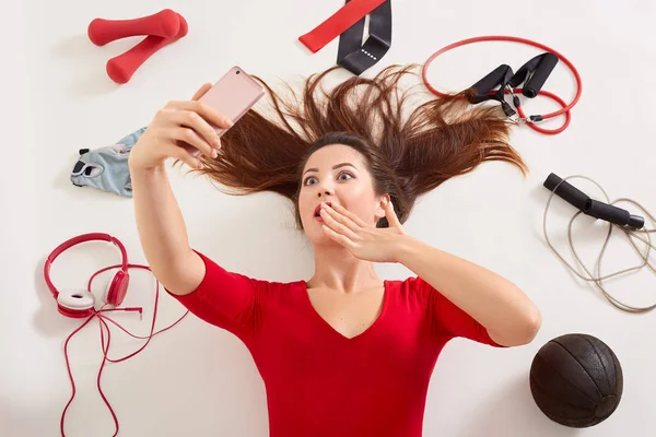 Sport woman laying on white surface with astonished facial expression, making selfie via her modern smart phone while covering mouth with hands, posing with different equipment needed for training. — Stock Photo, Image