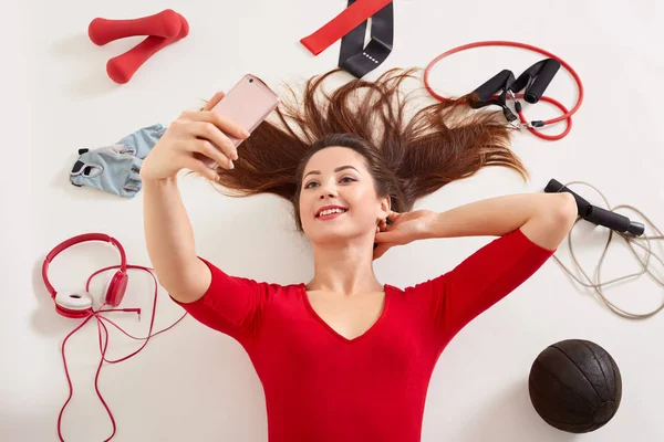 Sport woman smiling at camera of her moderm cell phone, making self picture at gym while laying on floor, young girl being at fitness center surrounded dumbbells, skipping rope, gloves, headphones. — Stock Photo, Image