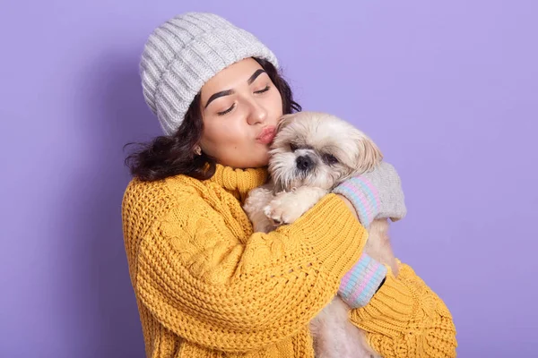 Chica de pelo oscuro con suéter amarillo cálido y gorra besando a su perrito, mujer europea dichosa expresando amor a su mascota favorita, señora mantiene los ojos cerrados, posando contra la pared del estudio lila . —  Fotos de Stock