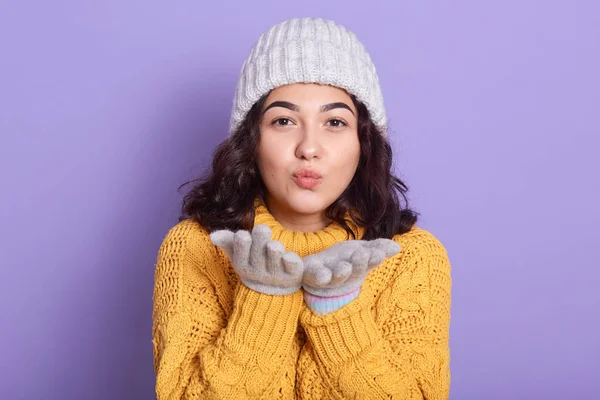 Mulher morena bonita veste camisola amarela quente, luvas de gorro branco enviando beijo de ar na câmera isolada sobre fundo lilás, fêmea com cabelo ondulado escuro e maquiagem leve, olhando para a câmera . — Fotografia de Stock