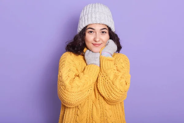 Gadis terinspirasi mengenakan sweater kuning yang nyaman, topi putih hangat dan sarung tangan, berpose dengan tampilan romantis di studio terhadap dinding ungu, wanita ceria terus tangan di bawah dagunya, mengekspresikan kebahagiaan . — Stok Foto