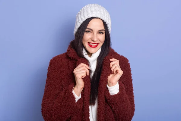Μοντέρνα γυναίκα με σκούρο καφέ παλτό και λευκό καπέλο χειμώνα ποζάρουν σε στούντιο απομονωμένο πάνω από το μπλε φόντο στούντιο, κοιτάζοντας κάμερα, έχει σκούρα μαλλιά και φωτεινό μακιγιάζ, κρατά τα χέρια στο γιακά του παλτού — Φωτογραφία Αρχείου