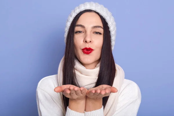 Wanita muda yang mengenakan pakaian musim dingin melakukan ciuman terbang dengan latar belakang biru, wanita dengan pomade merah, gadis berpakaian topi putih dan sweater, wanita cantik mengirimkan ciuman udara. Konsep orang . — Stok Foto