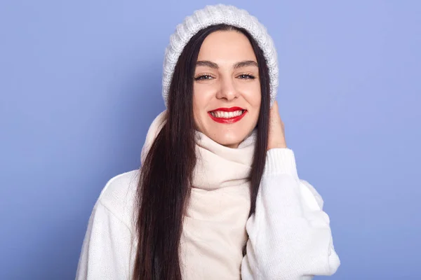 Retrato de hermosa mujer joven feliz con expresión facial feliz, chica con el pelo largo y oscuro y labios rojos. Chica sonriente lleva gorra blanca, bufanda y suéter. Fondo del estudio azul. Estilo de invierno . —  Fotos de Stock