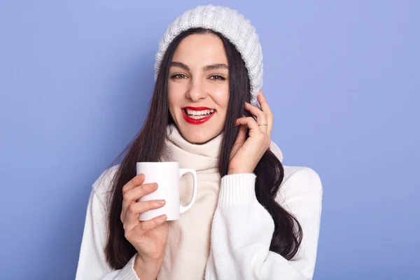 Молода приваблива жінка тримає кепку гарячого напою в руках, одягнена в білий теплий светр, шарф і кепка, позує ізольовано на синьому фоні, брюнетка з червоними губами, тримає руку біля обличчя — стокове фото