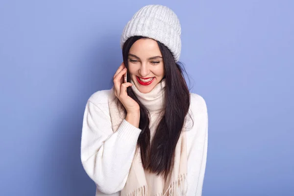 Happy young woman talking to her friend via smart phone isolated over blue background, lady with red lips dresses white warm clothes, brunette girl having joyful facial expression. People concept. — 스톡 사진